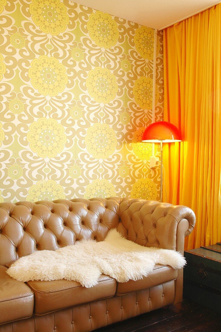 Weisses Tierfell auf brauner Ledercouch mit Zierknöpfen, in Zimmerecke Stehleuchte mit orangerotem Schirm vor Wand mit gemusterter Tapete im 70er Jahre Stil