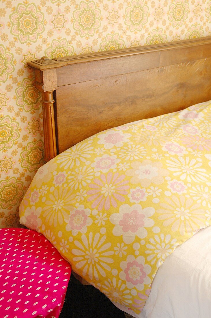 Ausschnitt eines Bettes mit Holz-Kopfteil, Blumenmuster auf Bettwäsche und Tapete an Wand