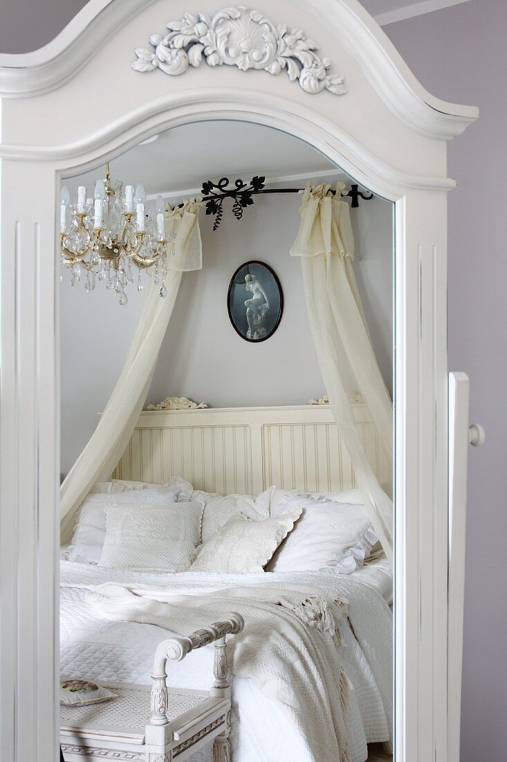 Mirror with white, carved wooden frame reflecting double bed with white bed linen and canopy