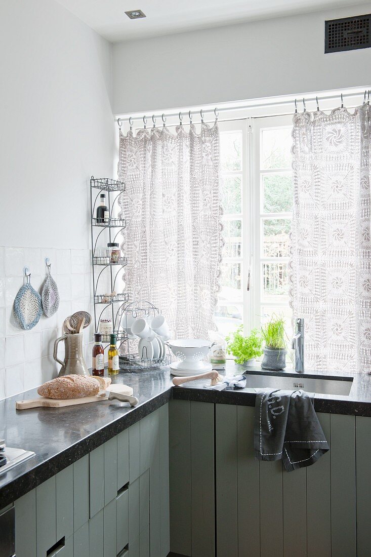 Ausschnitt einer Küche, Küchenzeile übereck mit dunkler Arbeitsplatte, und eingebautem Spülbecken, vor Fenster mit gehäkeltem Vorhang