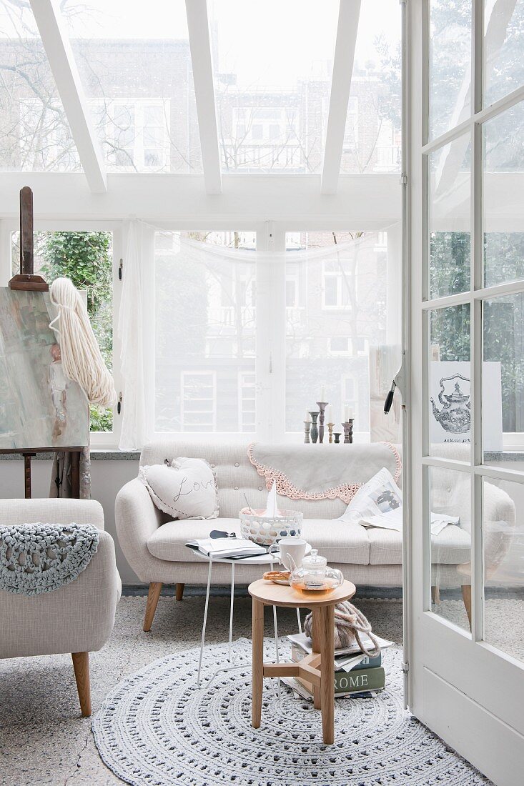 Blick durch offene Tür auf rundem Häkelteppich, Sessel und Sofa im Fiftiesstil in hellem Zimmer mit Verglasung