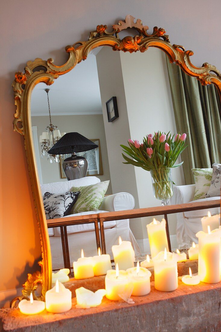 Lit, white pillar candles in front of mirror with antique gilt frame reflecting vase of tulips on coffee table