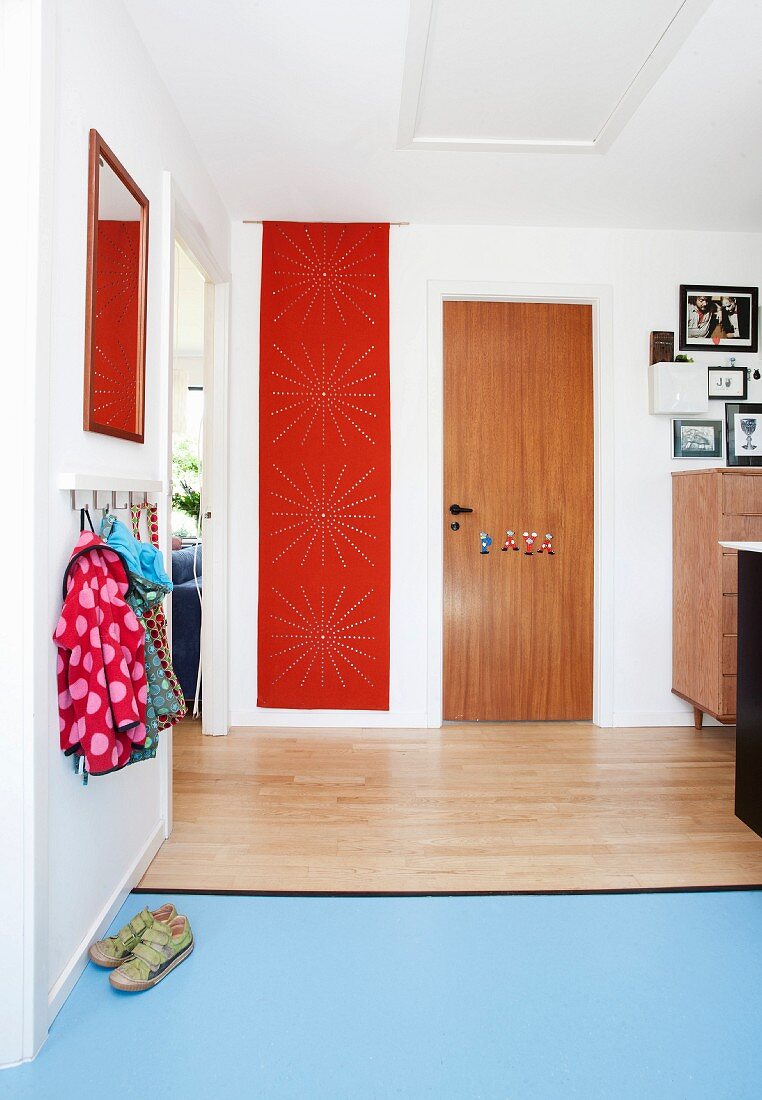 Gangbereich mit Kindergarderobe, verschiedene Beläge, Holz und hellblauer Linoleum auf Boden, im Hintergrund rote Fahne mit Lochmuster an Wand
