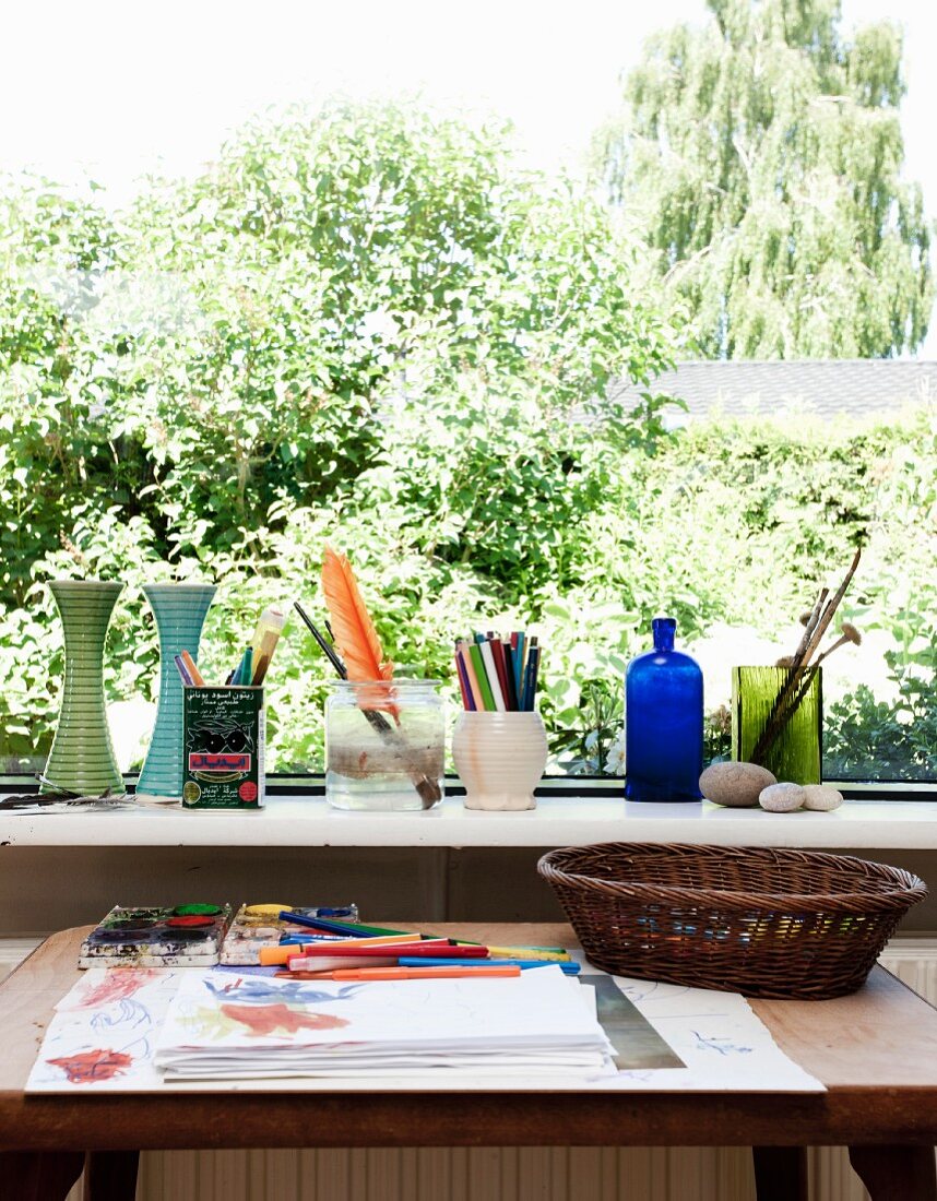 Tisch mit Malutensilien und Zeichnung vor Fenster mit Aussicht, auf Fensterbank Vasen und Behälter mit Stiften