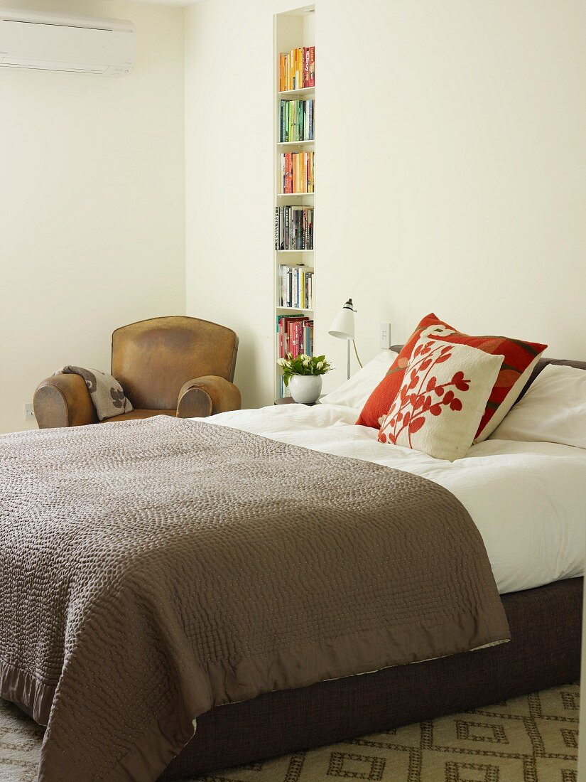 Boxspringbett mit brauner Tagesdecke, gemusterte Kissen in schlichtem Schlafzimmer, seitlich schmales Bücherregal in Wandnische eingebaut
