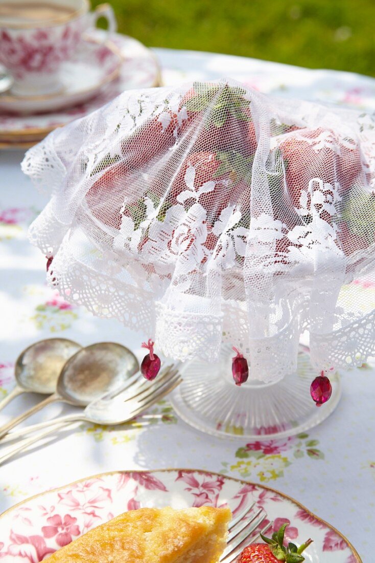 Selbstgenähte Fliegenschutzhaube aus weißem Spitzenstoff für Erdbeerschale auf romantisch gedecktem Gartentisch