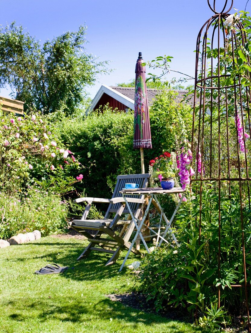 Holzstuhl und Klapptisch in sommerlichem Garten mit Vintage-Rankhilfe und Sonnenschirm