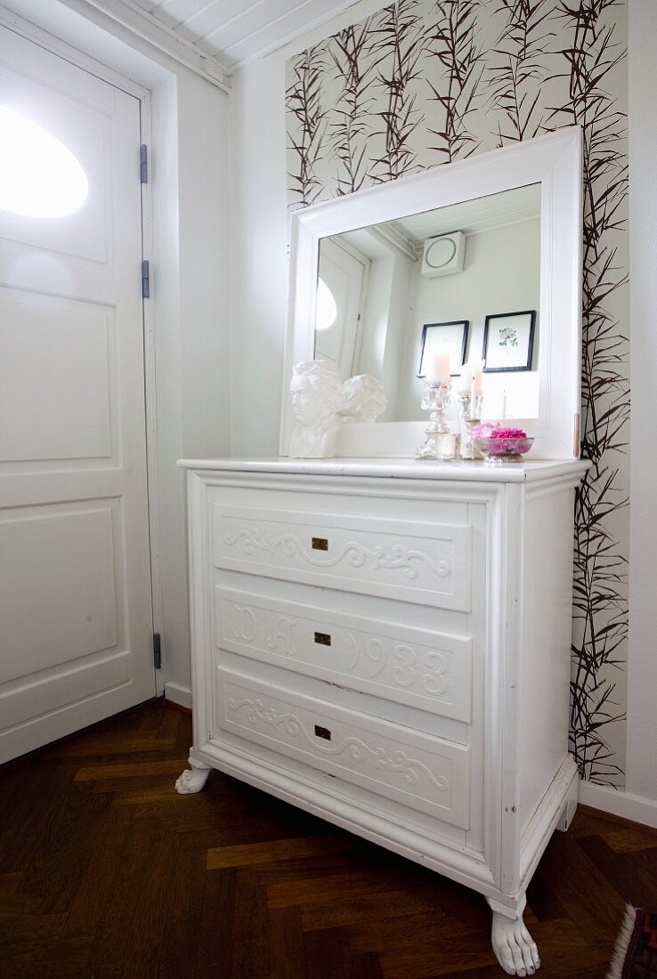 Weiß lackierte Kommode und gerahmter Spiegel vor tapezierter Wand mit Blattmuster