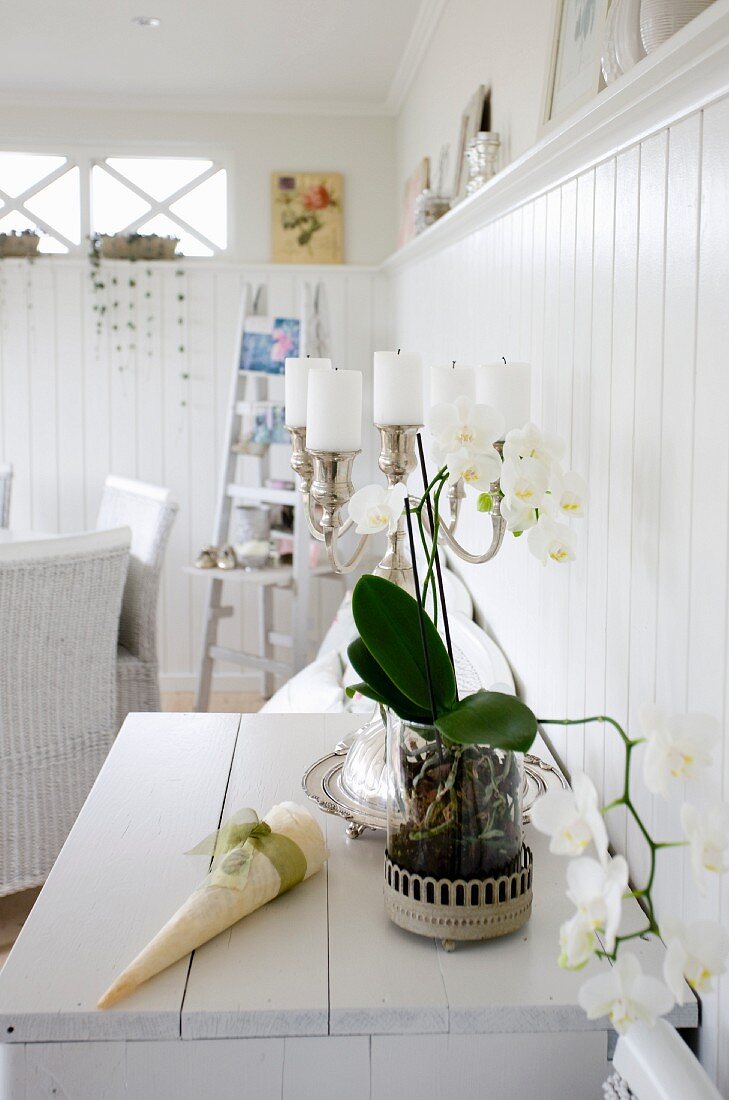 weiße Orchidee und mehrarmiger Kerzenhalter mit weissen Stumpenkerzen auf Kommode, vor weisser holzverkleideter Wand
