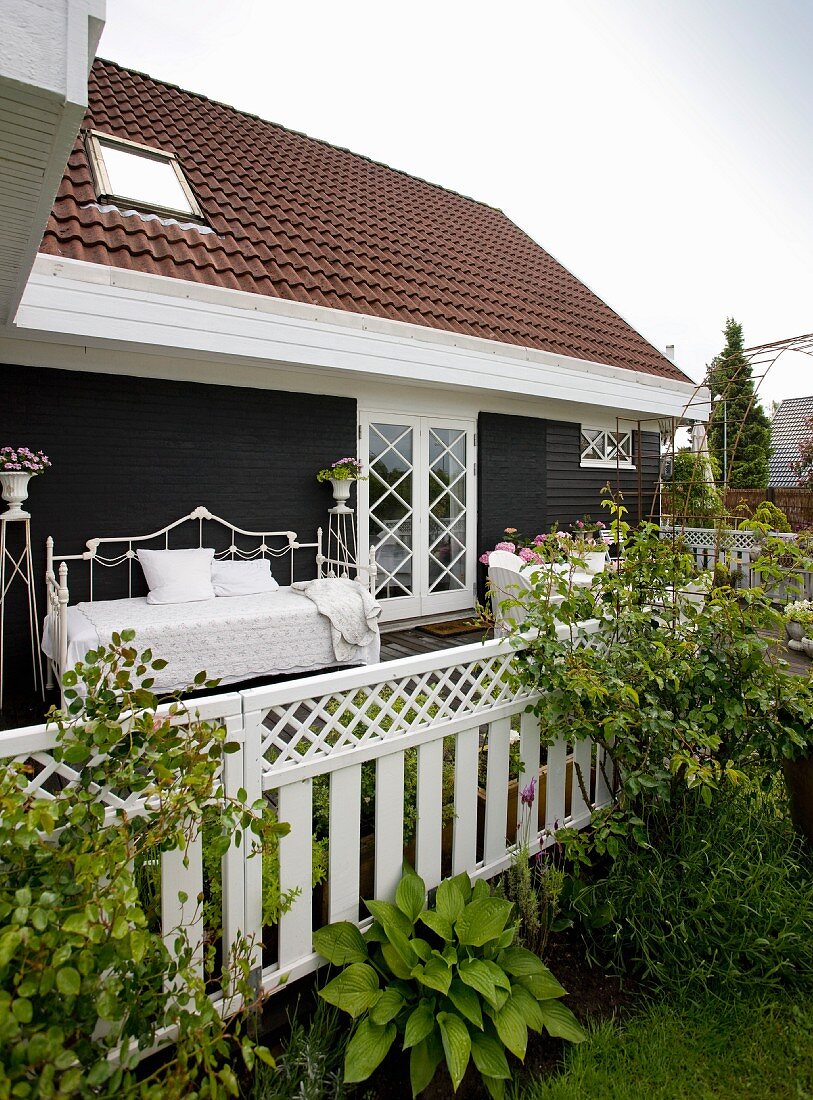 Blick von Garten auf Terrasse mit Bett aus weißem Metall vor dunkler Holzwand