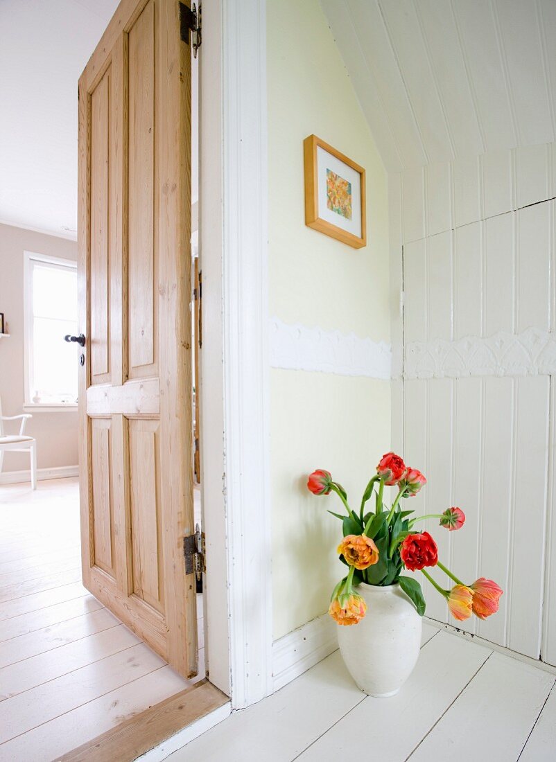 White vase of red and yellow tulips on white-painted wooden floor next to open interior door