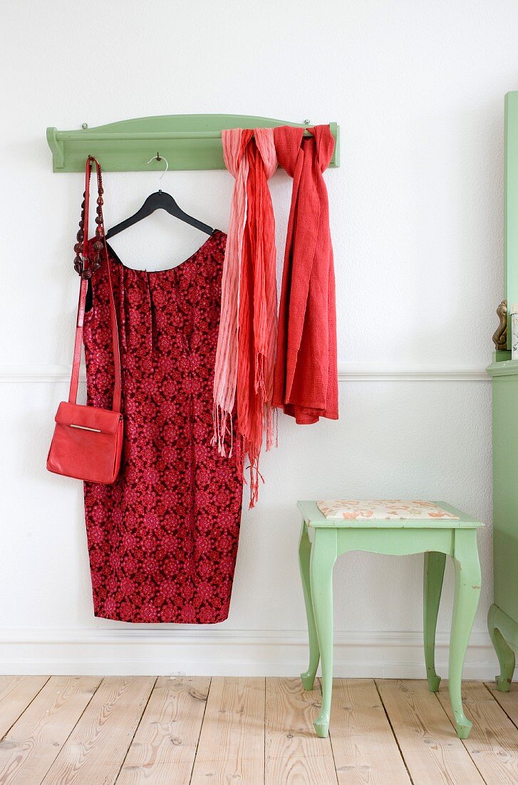Rotes Damenkleid auf Kleiderbügel und Tücher an grün lackierter Wandgarderobe, seitlich farblich passender Beistelltisch mit geschwungenen Beinen