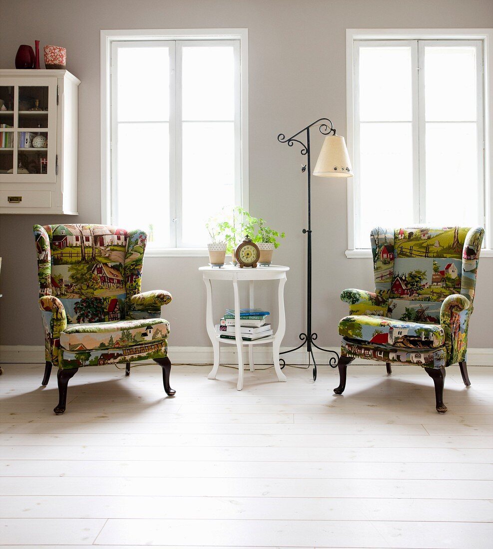 Weiß lackierter Beistelltisch und Stehleuchte mit schmiedeeisernem Gestell zwischen Sessel mit gemustertem Bezug, ländliche Motive, vor Fenster