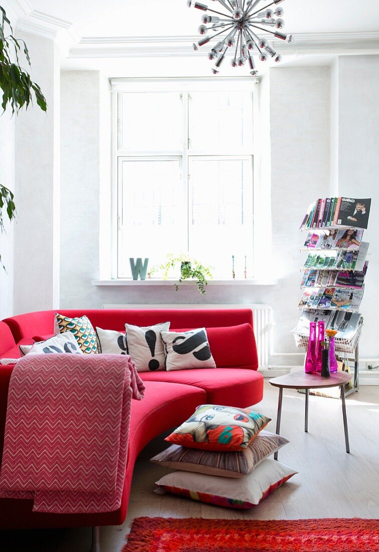 Rotes Designersofa mit Kissenstapel am Boden, Zeitschriftenständer und Beistelltisch mit Retroflair in renovierter Altbauwohnung