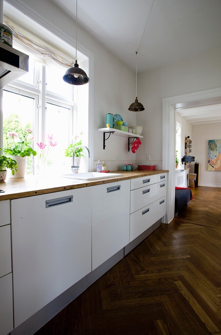 Kitchen counter with white base units below window in renovated period building with continuous herringbone parquet flooring