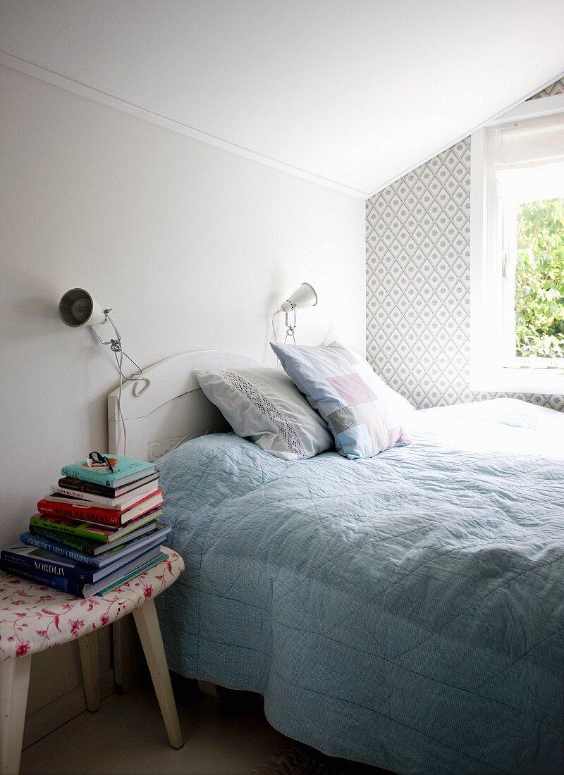 Doppelbett mit weißem Kopfteil am Fenster und Schemel als Nachttisch mit Bücherstapel, im Schlafzimmer