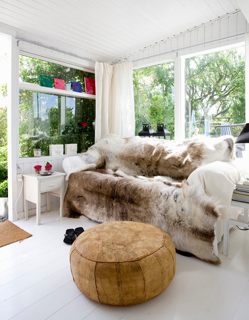 Gemütliches Sofa mit Rentierfell, davor Leder Sitzpouf in hellem Wintergarten