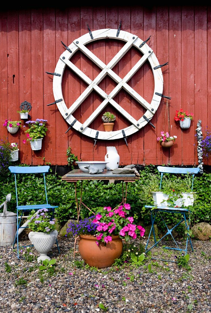 Altes Mühlenrad und Blumentöpfe an rotbrauner Holzwand, davor Gartenmöbel und blühende Blumen