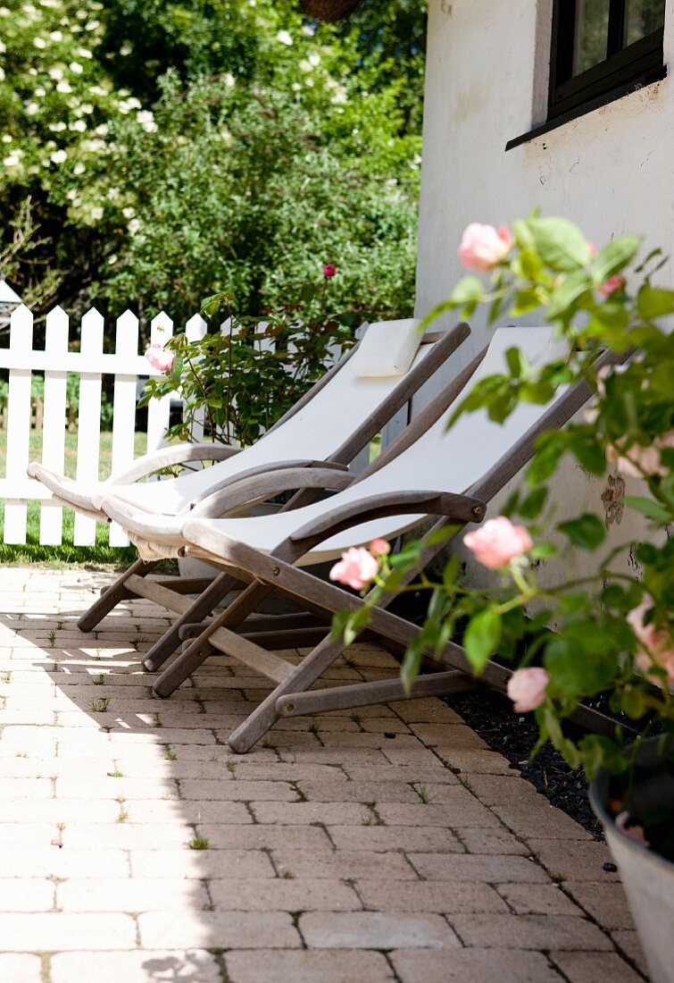 Holzliegestühle mit weißem Bezug auf Terrasse vor Wohnhaus, im Hintergrund Garten
