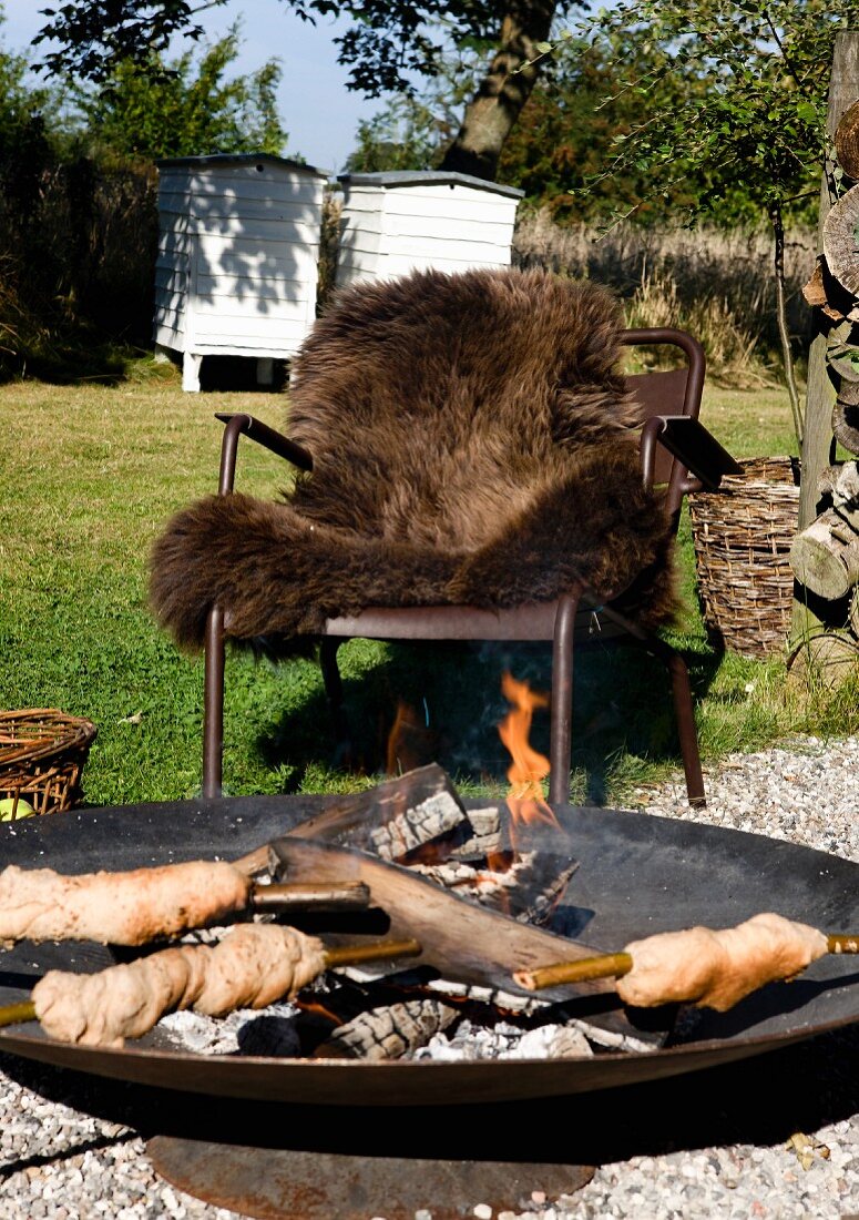 Campfire bread in fire bowl in front of outdoor armchair with brown fur blanket in garden