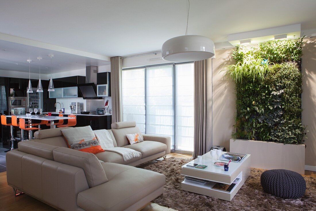 Loungebereich mit Überecksofa und Couchtisch vor vertikaler Pflanzendeko, im Hintergrund offene Küche mit orangefarbenen Barhockern