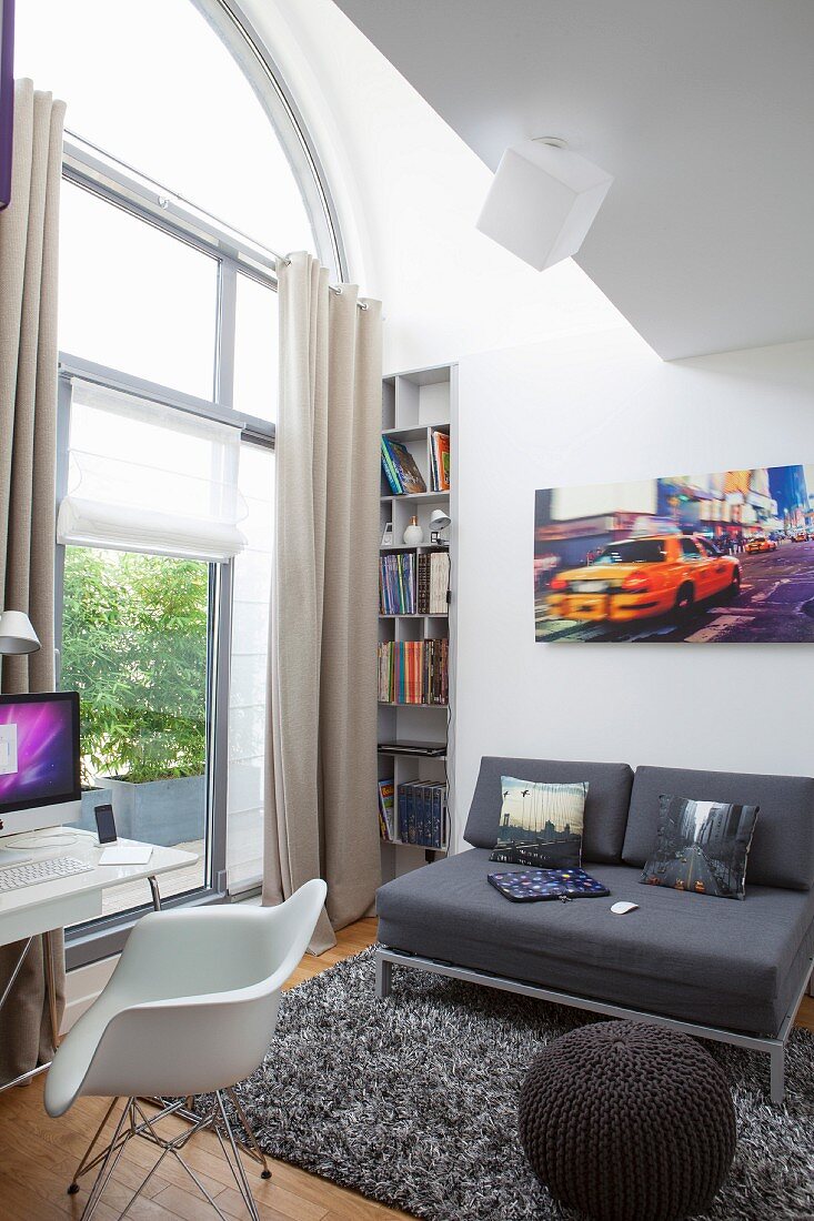 Sofa und Strick-Pouf auf Hochflorteppich vor hohem, Rundbogenfenster, vorne Schalenstuhl und Schreibtisch