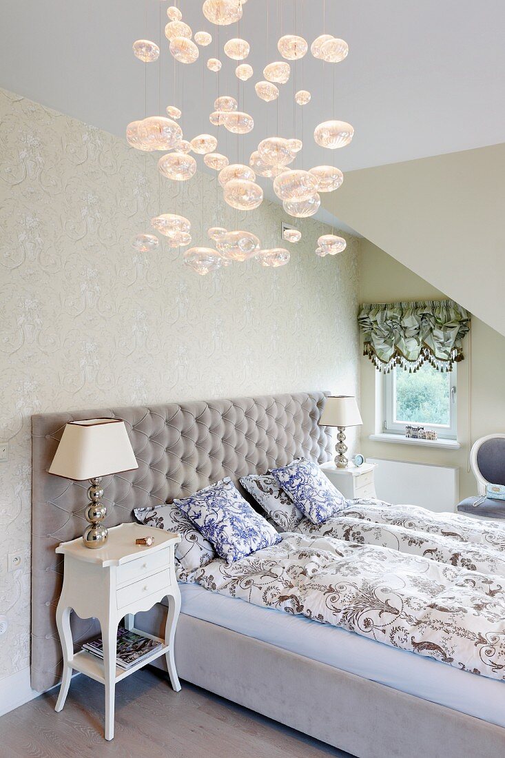Doppelbett mit gepolstertem Kopfteil und klassische Leuchten auf Antikkommoden in der Ecke eines Dachzimmers; Hängeleuchte mit Glasbubbles im Vordergrund