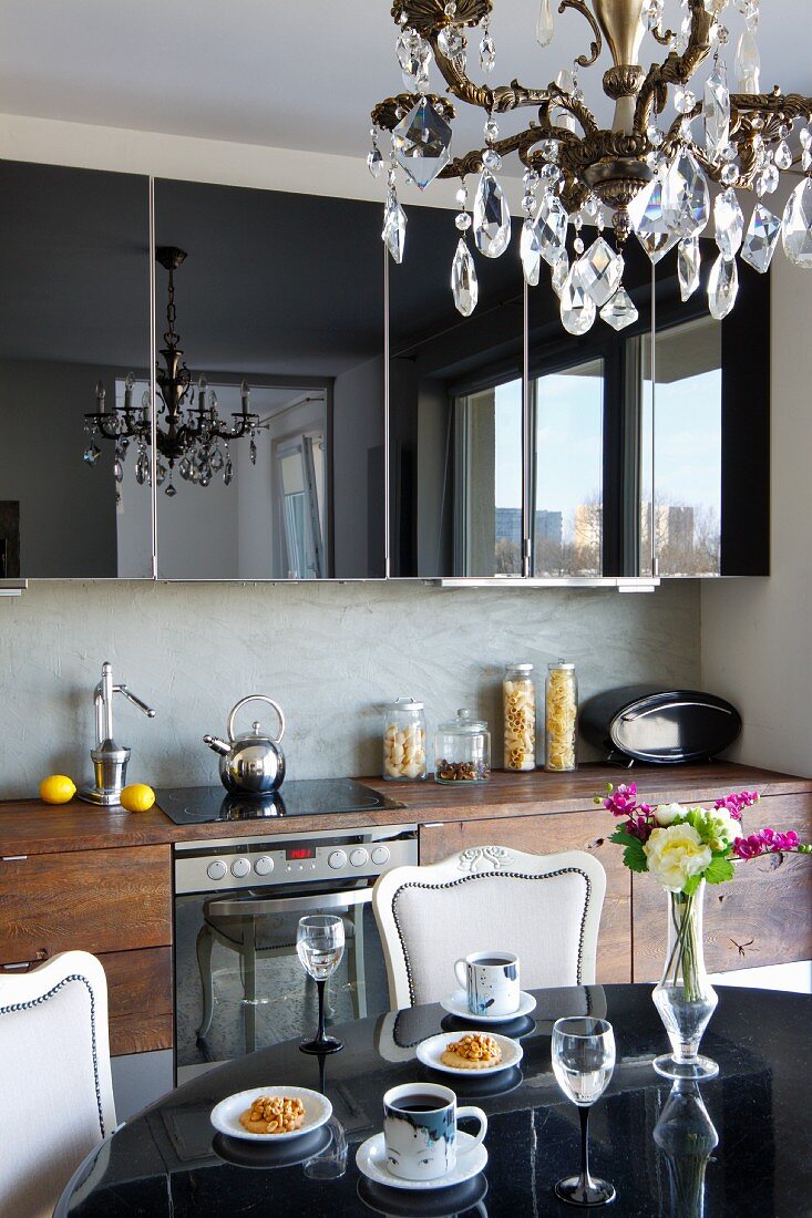 Kristallleuchter über elegantem Essplatz, dahinter moderne Küchenzeile mit hochglänzenden Oberschränken