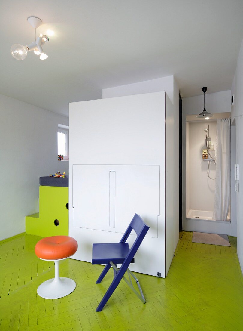 Retro stool and folding chair in front of closed modular kitchen on lime green parquet floor with shower cubicle in background