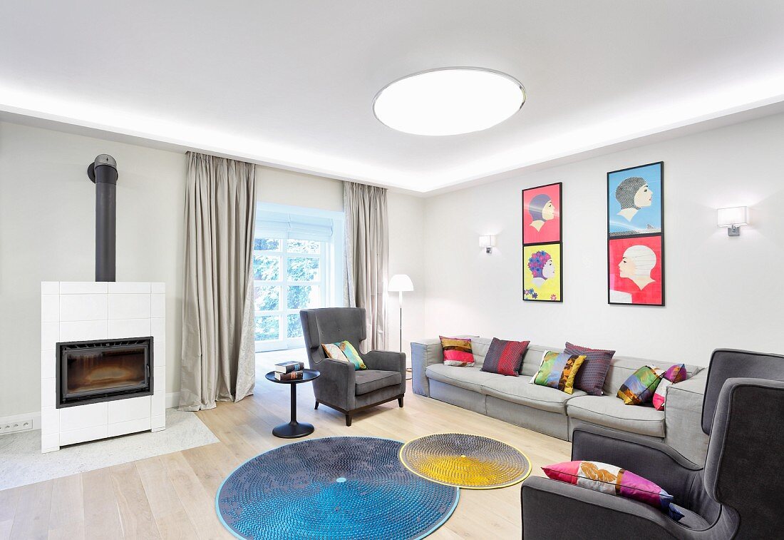 Bunte Kissen auf Sofa und Sessel in Grautönen, runde farbige Teppiche vor dem Kamin und Bilder im Pop-Art-Stil