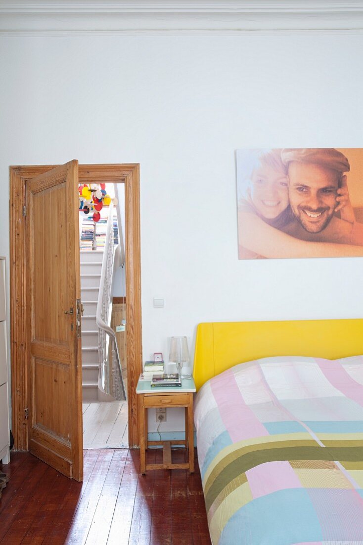 Doppelbett mit gelbem Kopfteil, Bettwäsche in Patchwork Muster, daneben kleiner Nachttisch und offene Tür mit Blick auf Treppe