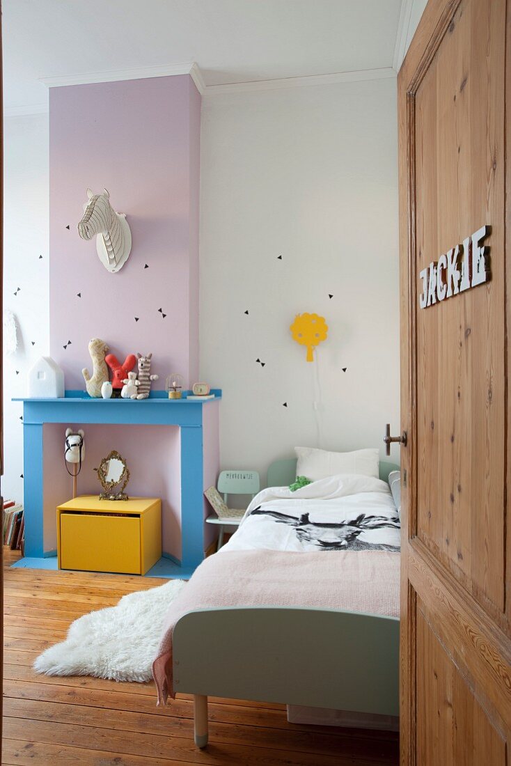 View through open door into child's bedroom; bed next to disused fireplace in bright colours below stylised animal head on wall