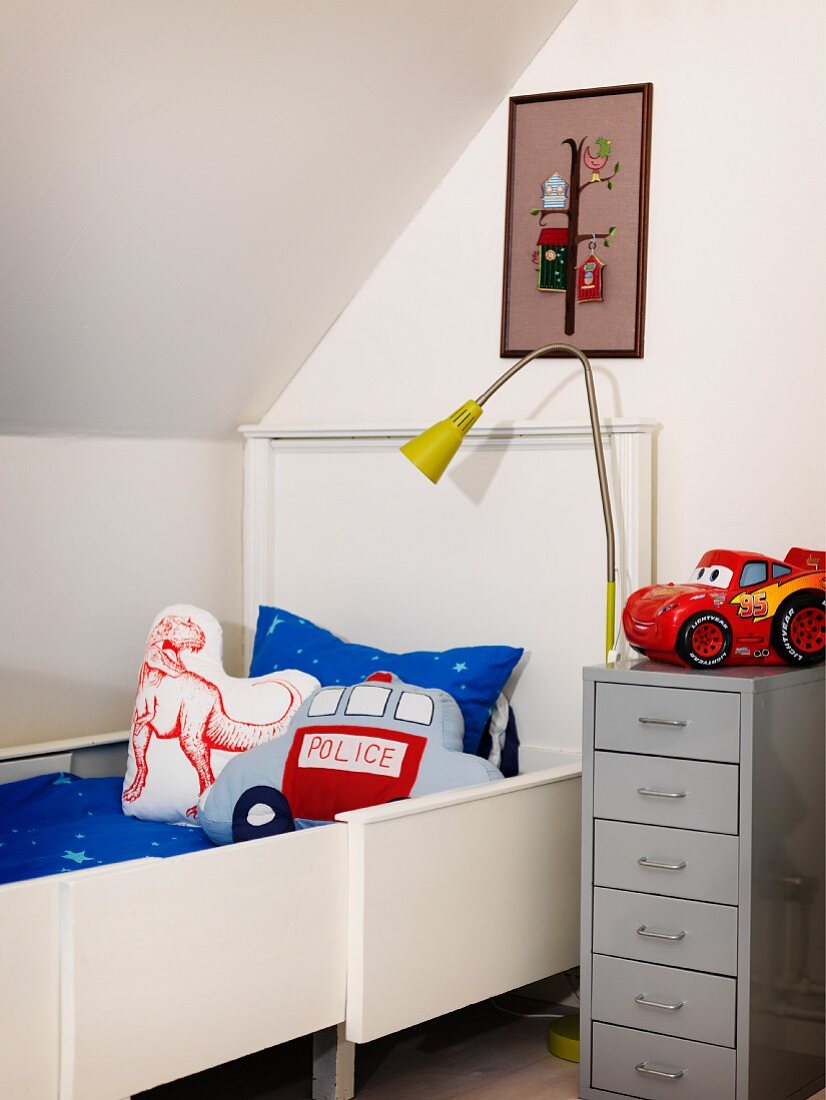 Jungenzimmer mit weißem ausziehbarem Bett und Dekokissen, grauem Metallschränkchen und Stehlampe