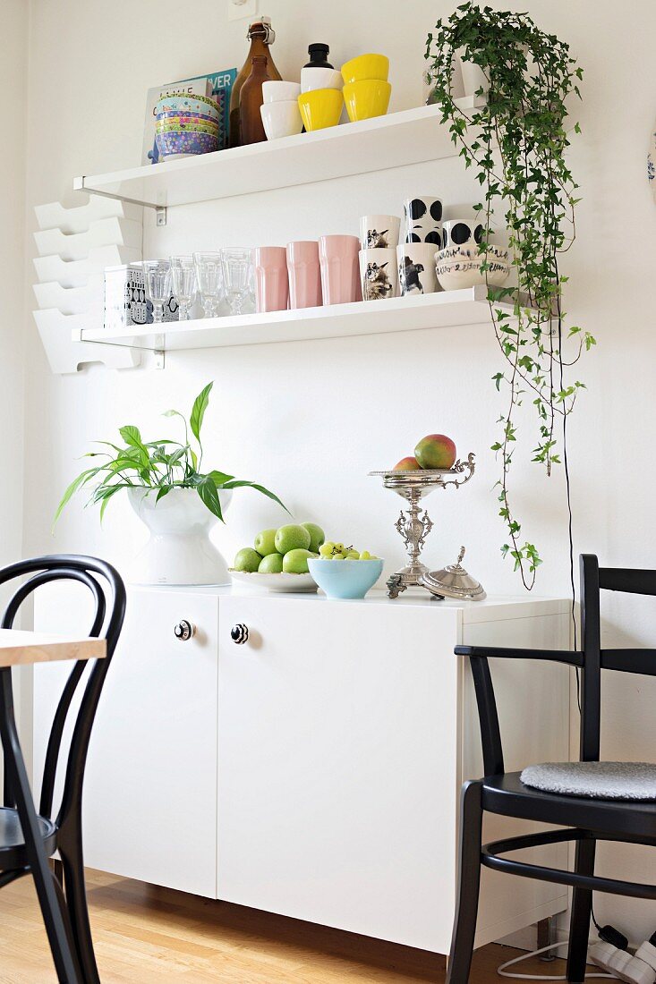 Fruit bowls and houseplants on white sideboard below white shelves on wall in dining room with black chairs