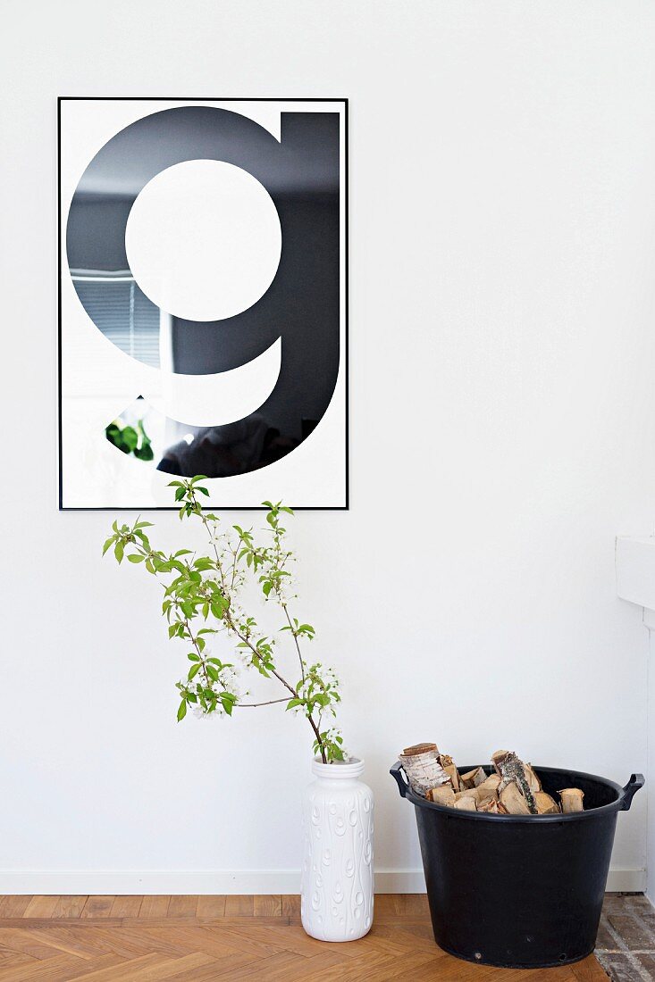 Framed, typographic poster of letter G on wall above leafy branch in white vase and bucket of logs on floor