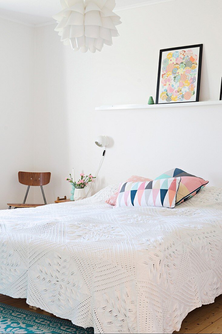 Doppelbett mit weisser Spitzen Tagesdecke, gemusterte Kissen, oberhalb an Wand weiße Ablage