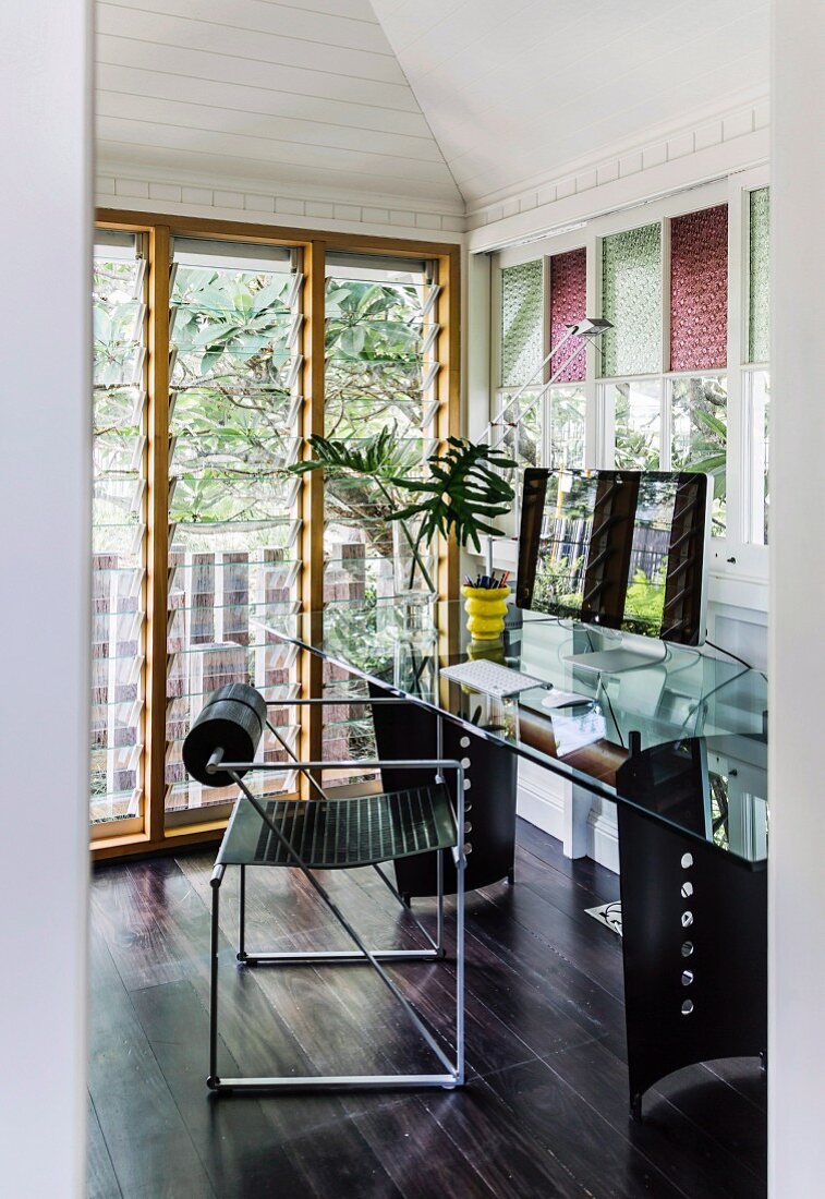 Moderner Stuhl aus Metall vor Schreibtisch mit Glasplatte, in Zimmerecke auf dunklem Dielenboden, seitlich Fenstertüren mit verstellbarem Lamellen Glas