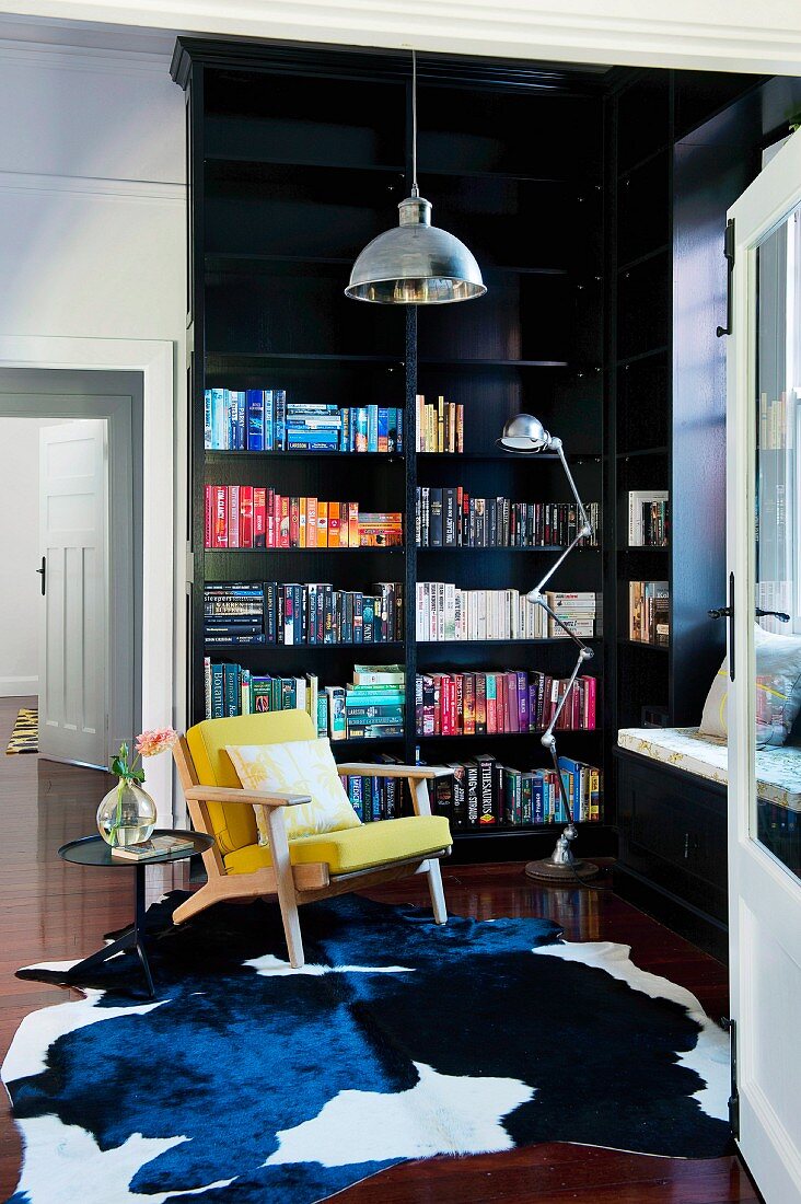 Schwarzes Einbaubücherregal mit farblich sortierten Büchern, Kuhfellteppich und senfgelbem Polstersessel in elegantem Altbauambiente
