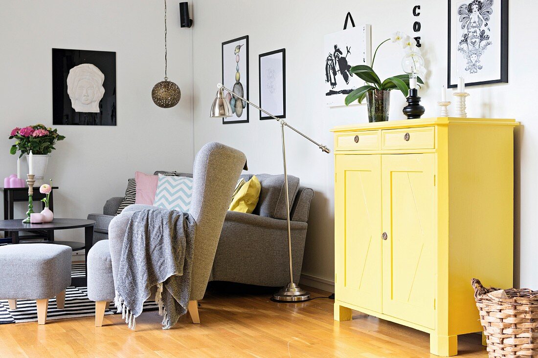 Yellow-painted cabinet next to seating area with grey armchair, matching footstool and retro-style chrome standard lamp