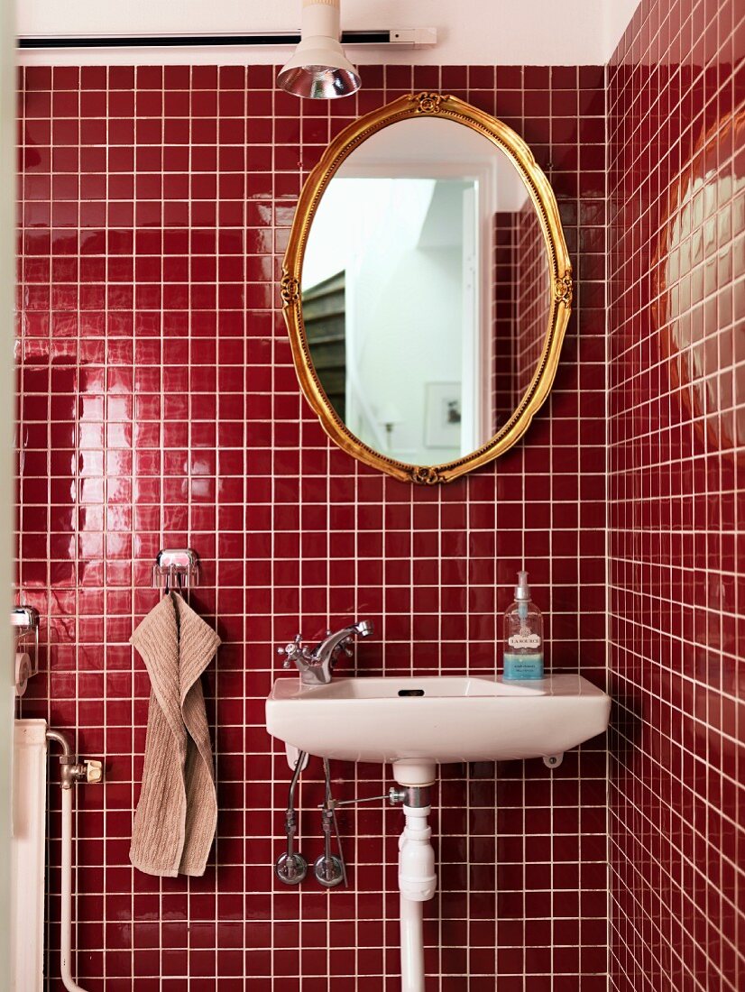 Sink below oval gilt-framed mirror on red-tiled wall in corner of modern bathroom