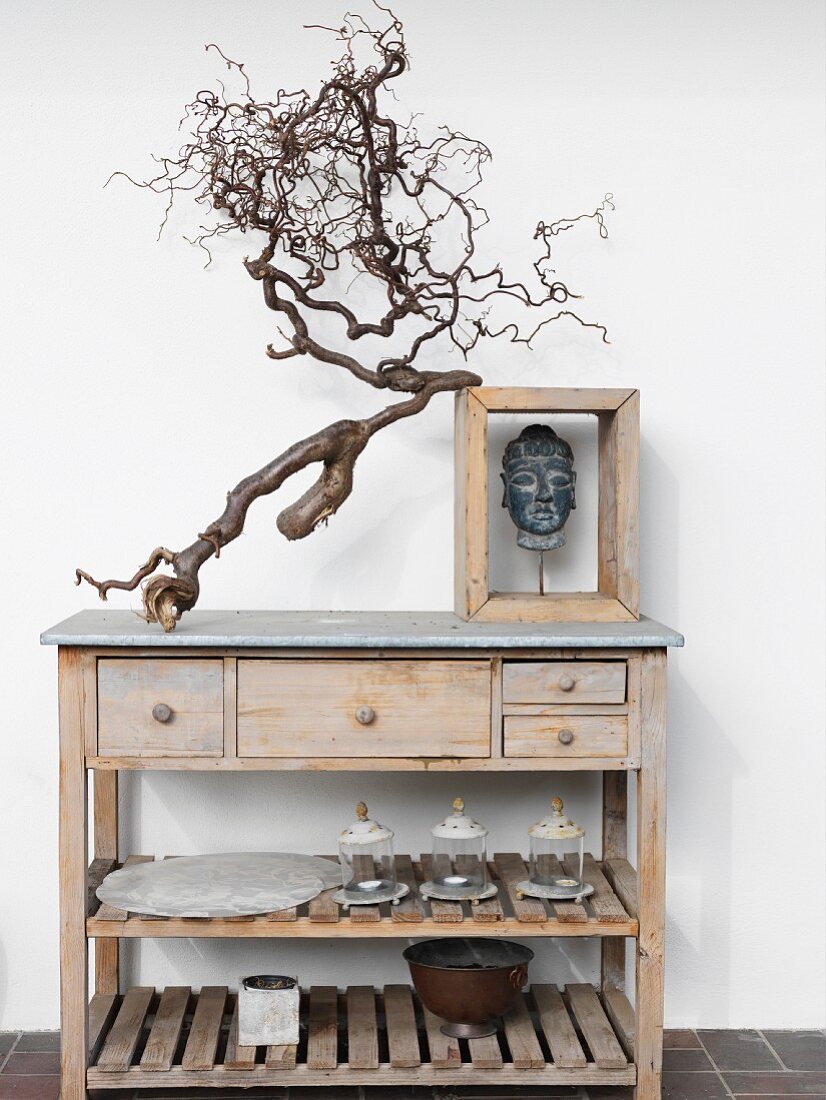 Gnarled branch and head of Buddha in wooden frame on top of simple wooden sideboard with drawers and shelves