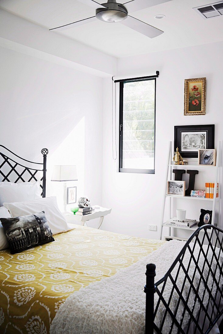 Yellow floral bedspread on black metal bed in front of small ladder-style shelves of ornaments