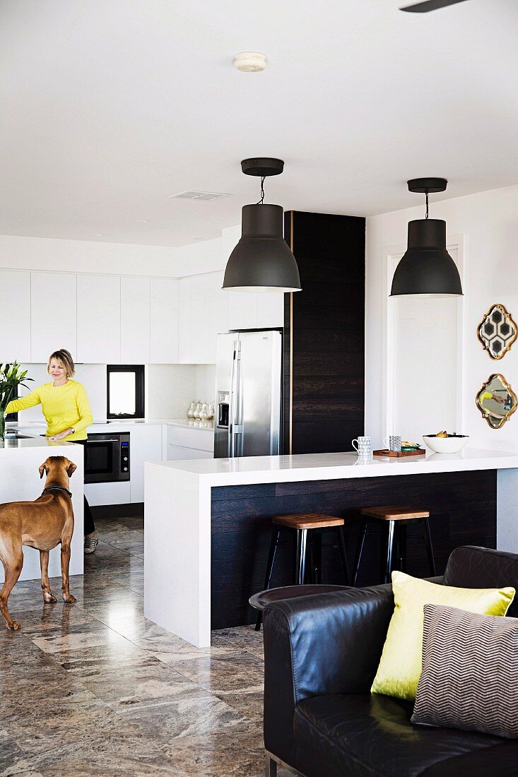 Woman and dog in open-plan fitted kitchen with striking pendant lamps above kitchen counter and retro stools; leather armchair with scatter cushions in foreground