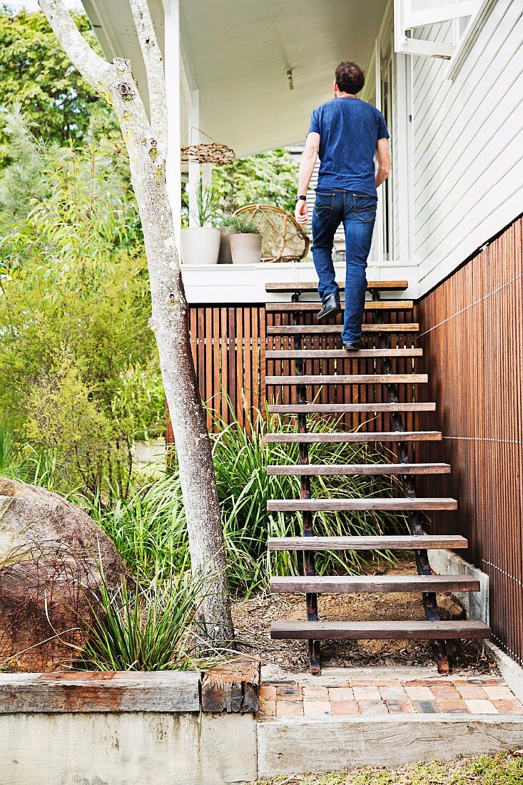 Mann geht geländerlose rustikale Holztreppe zum Hauseingang hoch, grüner Vorgarten mit Gräsern und Büschen