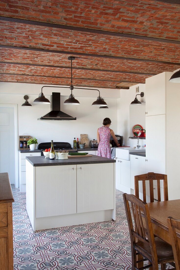 Schlichte Einbauküche mit freistehendem Block auf gemustertem Fliesenboden, im Hintergrund Frau