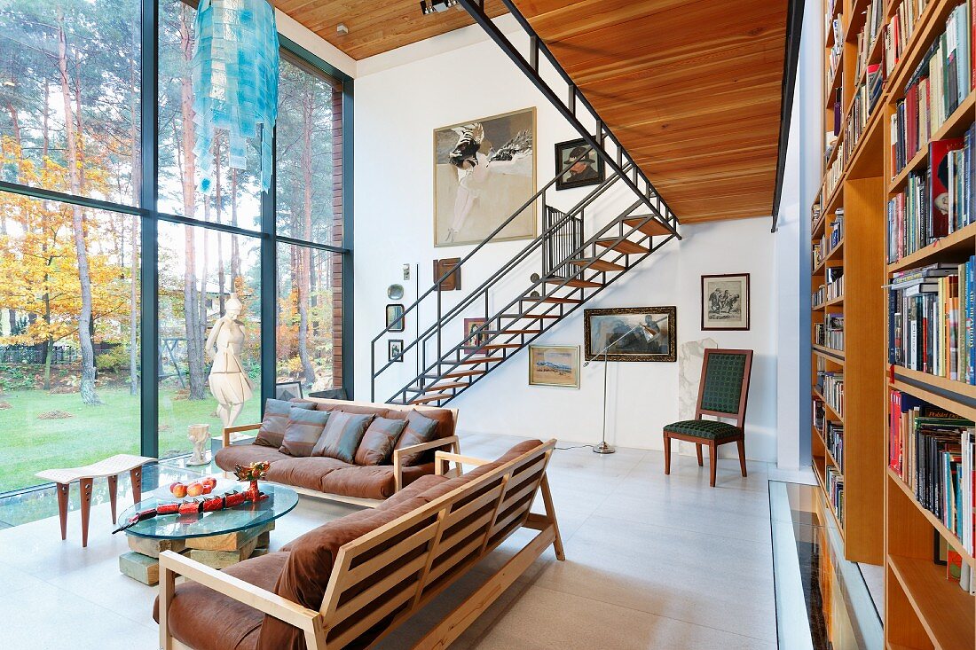 Sofagestelle aus Holz mit Lederkissen und Beistelltische vor zweigeschossiger Fensterfront, gegenüber Bücherregal unter Galerie mit leichter Stahlholztreppe