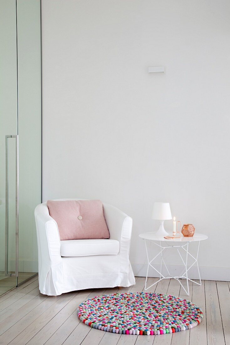 Colourful, round pompom rug in front of loose-covered armchair and delicate side table