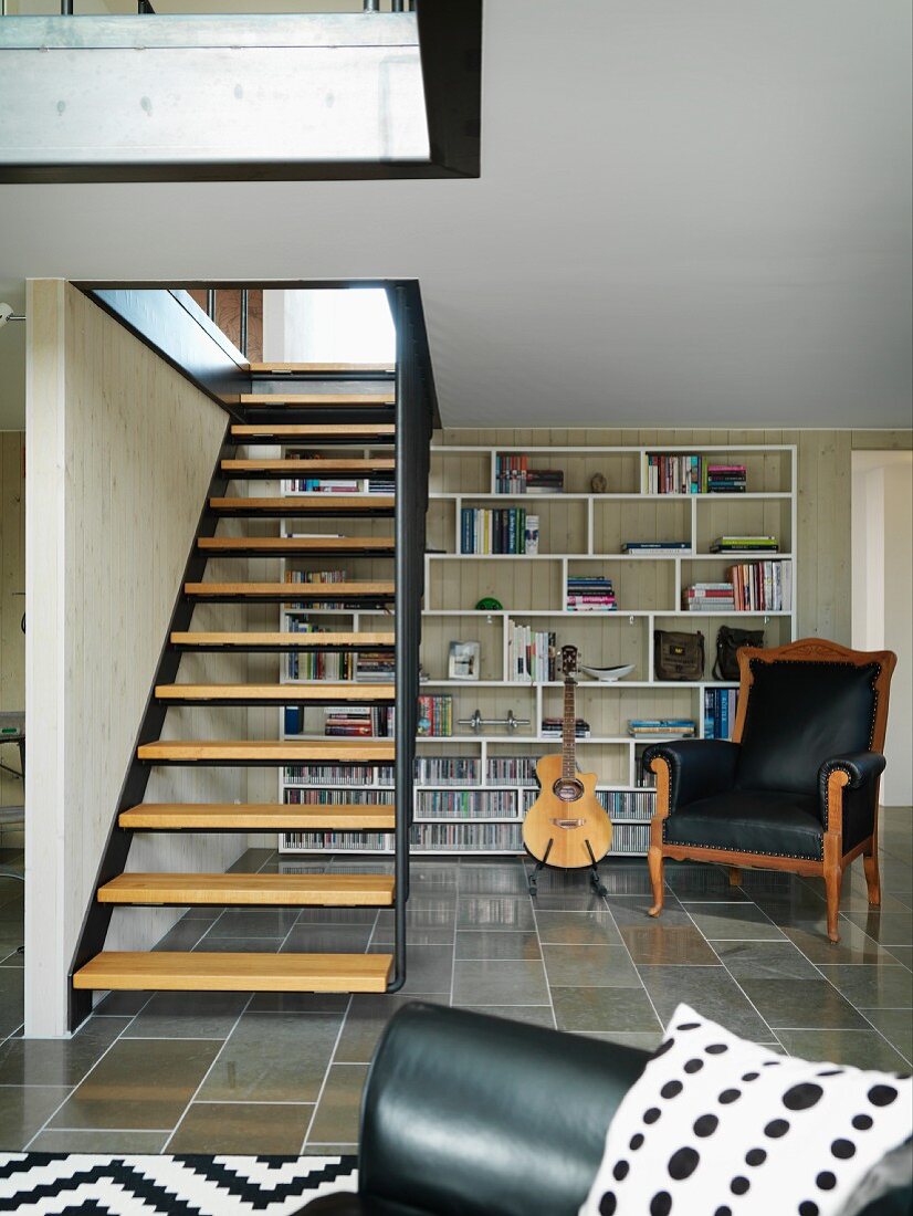 Treppe aus Metall mit Holztrittstufen im Wohnzimmer, im Hintergrund Sessel mit schwarzem Lederbezug vor modernem Wandregal