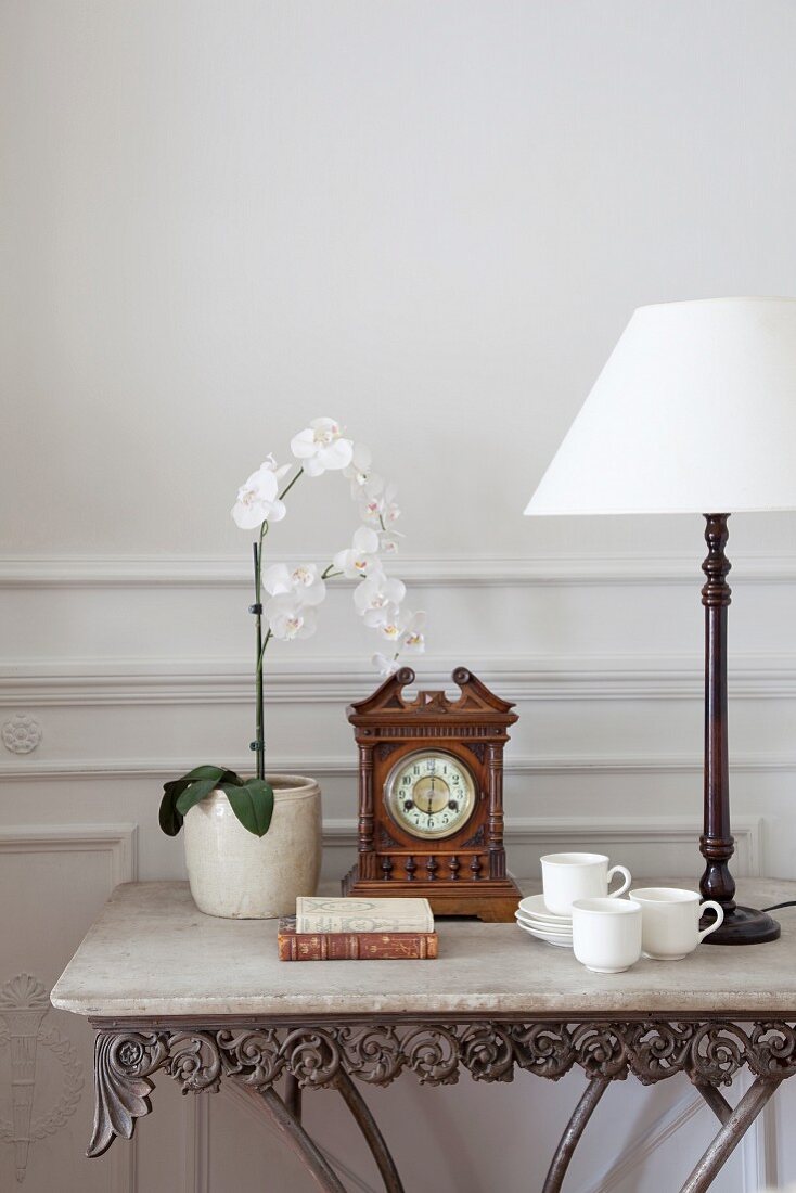 Orchid, antique table clock and table lamp with white lampshade on antique console table with stone top