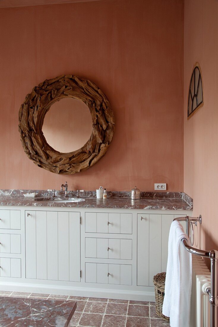 Detail of washstand with wooden front painted pale grey below round mirror with wooden frame on pink-glazed wall